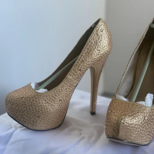 Guldglittriga pumps med platå - Snygga pumps i guld med glittrande stenar över hela skon. Skorna har hög klack och platåsula framtill för extra höjd och attityd. Perfekta för dig som vill sticka ut och glänsa. Klassisk rund tå och elegant form.