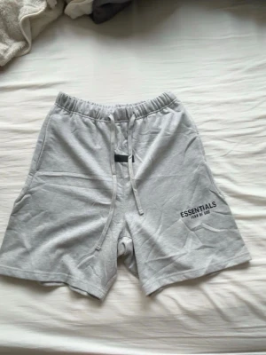 Grå Essentials Fear of God shorts - Snygga ljusgrå shorts från Essentials Fear of God med elastisk midja och snörning. Mjuk bomullskänsla och tryckt logga på vänster ben. Perfekta för chill dagar eller streetwear-stil.
