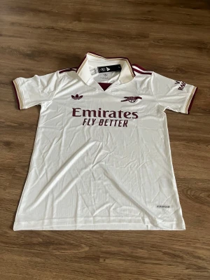 Arsenal vit matchtröja Adidas - Snygg vit Arsenal matchtröja från Adidas med vinröda detaljer och klubbmärke på bröstet. Tröjan har klassisk krage, korta ärmar och 'Emirates Fly Better' tryckt på framsidan. Tillverkad i lätt och ventilerande material (AEROREADY). Perfekt för fotboll eller träning.