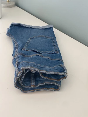 Blå jeansshorts med uppvik - Snygga blå jeansshorts med uppvikta benslut och klassisk femficksdesign. Shortsen har knapp och dragkedja framtill samt kontrastsömmar. Perfekta för varma dagar och enkla att matcha med olika toppar.