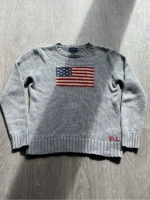 Grå stickad tröja Polo Ralph Lauren - Snygg grå stickad tröja från Polo Ralph Lauren med amerikansk flagga på bröstet och RL-broderi i rött vid nederkanten. Tröjan har rund halsringning, ribbstickade muddar och är perfekt för dig som gillar klassisk college-stil.