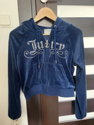 Blå hoodie från Juicy Couture - Mjuk och lyxig blå velour hoodie från Juicy Couture med dragkedja framtill och stor ficka. Snyggt Juicy-tryck i strass på bröstet och huva med dragsko. Perfekt för en avslappnad och trendig look.