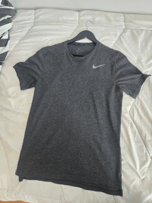 Mörkgrå Nike t-shirt - Nyskick Snygg mörkgrå t-shirt från Nike med klassisk logga på bröstet. Tillverkad i ett mjukt och funktionellt material som andas, perfekt för träning. T-shirten har rund halsringning och korta ärmar för en sportig look.
