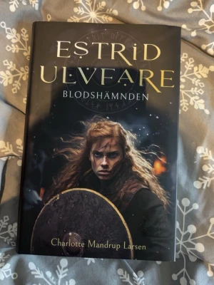 Estrid Ulvfare: Blodshämnden - Säljer boken 'Estrid Ulvfare: Blodshämnden' av Charlotte Mandrup Larsen. En svart hårdinbunden roman med guldfärgad text på omslaget och ett dramatiskt vikingamotiv. Första delen i en serie om vikingakvinnan Estrid, fylld av äventyr, mytologi och strid.