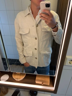 Beige overshirt från H&M - Stilren beige overshirt från H&M med fyra stora fickor framtill och knäppning med knappar. Tillverkad i ett lätt bomullstyg, perfekt för lager på lager. Klassisk krage och avslappnad passform som funkar till jeans eller chinos.