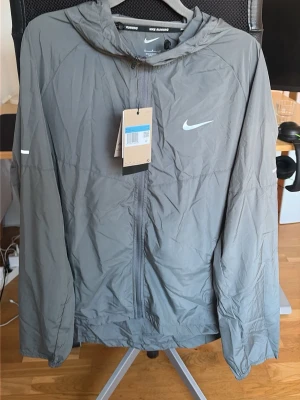Grå Nike miler jacka  - Snygg grå vindjacka från Nike med huva och dragkedja framtill. Jackan har reflekterande detaljer och Nike-logga på bröstet. Tillverkade i lätt, vindtätt material som passar perfekt för löpning eller promenader. Enkel och stilren design. PRIS KAN DISKUTERAS!
