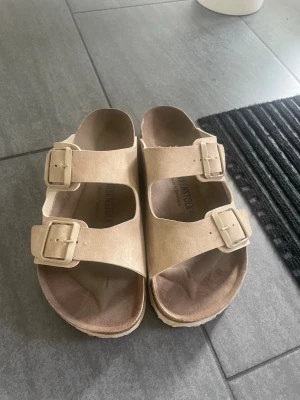 Beige sandaler från Birkenstock - Snygga beige sandaler från Birkenstock med två breda remmar och justerbara spännen i metall. Tillverkade i mockaliknande material med bekväm fotbädd och räfflad sula. Perfekta för en avslappnad och trendig stil.