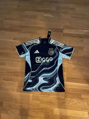 Ajax x Adidas matchtröja blå mönstrad - Snygg blå matchtröja från Ajax i samarbete med Adidas. Tröjan har ett unikt vågigt mönster i olika blå toner, vita detaljer och tre ränder på axlarna. Ajax klubbmärke och Ziggo-logga på bröstet. Perfekt för fotbollsfans!     Fraktar samma eller dagen efter du köpt. 
