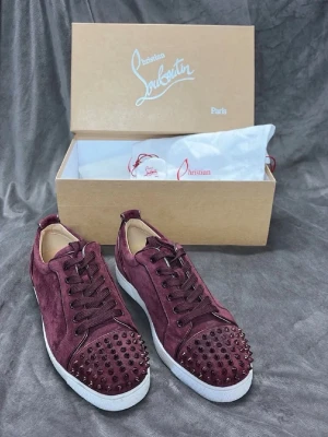 Vinröda sneakers från Christian Louboutin - Snygga vinröda sneakers i mocka från Christian Louboutin med ikoniska nitar på tån och vita sulor. Skorna har matchande vinröda skosnören och en lyxig känsla med detaljer i metall. Perfekta för dig som vill sticka ut med en exklusiv look.