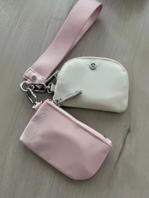 Lululemon Dual Pouch Wristlet rosa/vit - Snygg och praktisk wristlet från Lululemon med två små plånböcker i rosa och vitt. Båda facken har dragkedja och sitter fast i en bred handledsrem. Perfekt för kort, mynt och småprylar. Tillverkad i slitstarkt tyg med metall-detaljer och Lululemon-logga.