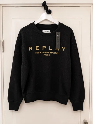 Svart sweatshirt från Replay - Svart sweatshirt från Replay med guldfärgad text 'REPLAY RUE ETIENNE MARCEL PARIS' på bröstet. Klassisk rund halsringning och ribbade muddar vid ärmslut och nederkant. Tillverkad i mjukt material som passar perfekt till en avslappnad stil.
