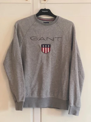 Grå sweatshirt från GANT - Klassisk grå sweatshirt från GANT med rund halsringning och broderad logga samt sköld med stjärnor och ränder på bröstet. Tröjan har ribbade muddar vid ärmslut och nederkant. Perfekt till ett avslappnat streetwear-inspirerat uttryck.