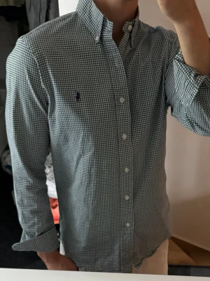 Grönvitrutig skjorta från Ralph Lauren - Klassisk grönvitrutig skjorta från Ralph Lauren med button-down krage och lång ärm. Skjortan har vita knappar framtill och en liten broderad logga på bröstet. Tillverkad i mjuk bomull som känns skön mot huden. Perfekt för en clean och stilren look. Lite missfärgad i kragen annars bra skick 