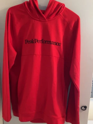Röd hoodie från Peak Performance - Snygg röd hoodie från Peak Performance med svart logotyp broderad på bröstet. Hoodien har huva och långa ärmar, samt en klassisk passform. Perfekt för dig som gillar sportig och stilren design. Materialet känns mjukt och bekvämt.