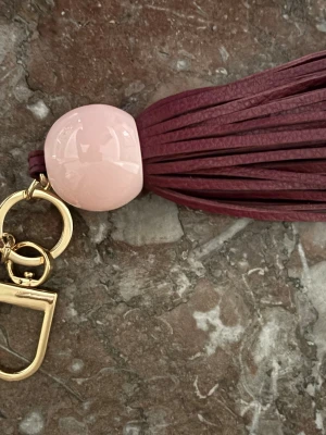 Tory Burch väsksmycke / bag charm med lädertofs – vinröd & rosa, originalask - Säljer ett supervackert Tory Burch väsksmycke / bag charm / nyckelring med elegant vinröd lädertofs, ljusrosa blank resin-detalj och guldfärgade metalldetaljer med den klassiska Tory Burch Double T-loggan.  Den är perfekt att använda som väsksmycke på handväska, tote bag eller nycklar – en liten detalj som direkt gör väskan mer lyxig och genomtänkt. Färgerna är väldigt fina ihop: burgundy/vinrött, blush pink och guld, vilket ger en exklusiv, feminin och trendig “designer accessory”-känsla utan a