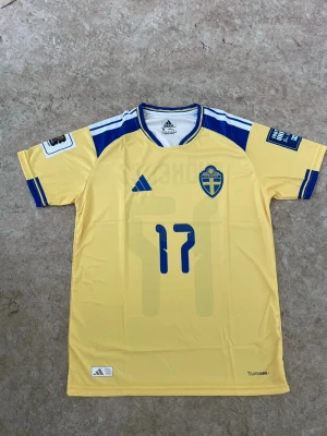 Svensk landslagströja Adidas nr 17 - Gul svensk landslagströja från Adidas med blå detaljer och nummer 17 på både fram- och baksidan. Tröjan har namntrycket 'Gyökeres' på ryggen, blåa ränder på axlarna och ärmarna samt broderad landslagssköld på bröstet. Tillverkad i lätt och ventilerande material.