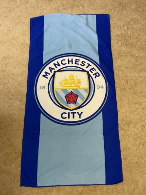 Manchester City strandhandduk blå - Stor strandhandduk i blått med Manchester Citys klubbmärke tryckt i mitten. Handduken har breda ränder i ljusblått och mörkblått och är tillverkad i mjukt material, perfekt för stranden eller poolen för alla City-fans.