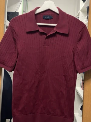 Vinröd ribbad polotopp från RILEY - Säljer en vinröd ribbad polotopp från RILEY med korta ärmar och klassisk krage. Har en snygg struktur och är tillverkad i ett mjukt material. Passar S/M
