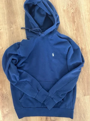 Ralph Lauren Hoodie - Snygg blå hoodie från Ralph Lauren med klassisk broderad logga på bröstet. Hoodien har huva med dragsko och en stor magficka. Tillverkad i mjukt bomullsmaterial som känns skönt mot huden. Perfekt för en avslappnad och stilren look. Enda fel är litet slitage vid tröjans ficka. Skriv för frågor eller funderingar 🙌 Tröjan är självklart äkta