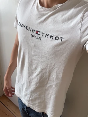 Vit Tommy Hilfiger t-shirt - Vit t-shirt från Tommy Hilfiger i slim fit-modell, tillverkad av ekologisk bomull. Framsidan har ett tryck med märkesnamnet och loggan samt texten 'Est. 1985'. Klassisk rund halsringning och korta ärmar.