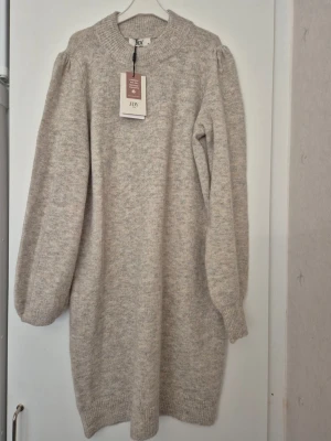 Klänning - Mysig beige stickad klänning från JDY med lång ärm och ribbade muddar. Klänningen har en rak passform och mjukt material som känns skönt mot huden. Perfekt för dig som gillar en enkel och stilren look.
