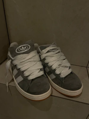 Grå Adidas sneakers med vita ränder - Snygga grå sneakers från Adidas med klassiska vita ränder på sidorna och stor vit logga på plösen. Skorna har breda vita skosnören och en gummisula med brun undersida. Ovandelen är i mocka och designen är stilren och sportig, perfekt för en streetwear-look.