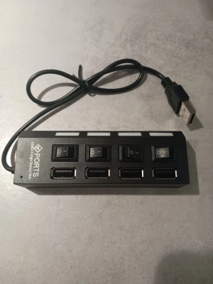 Svart USB-hubb med 4 portar - Praktisk USB-hubb med fyra USB-portar och individuella strömbrytare för varje port. Hubben är svart och har en rektangulär form med en USB-kabel för anslutning till dator eller annan enhet. Perfekt för att koppla in flera USB-enheter samtidigt.
