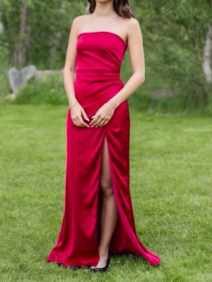Röd balklänning i satin med slits - Säljer en elegant röd balklänning i glansigt satinmaterial. Klänningen har en hög slits framtill och en figurnära passform som ger en snygg siluett. Perfekt för dig som vill sticka ut med en stilren och trendig look. Har själv sytt på en liten knapp vid slitsen för att den var väldigt lång, ( går att ta bort den om man vill ha högre slits). Klänningen är köpt från Nelly och endast använd 2 gånger. Inga defekter på klänningen. Köpt för 800kr
