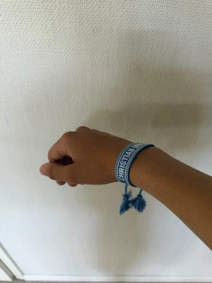 Dior armband - Snyggt blått tygarmband från Christian Dior med vit text och dekorativa tofsar i ändarna. Armbandet har ett vävt mönster och justerbar knytning för perfekt passform. Perfekt accessoar för att lyfta din outfit med en exklusiv touch.