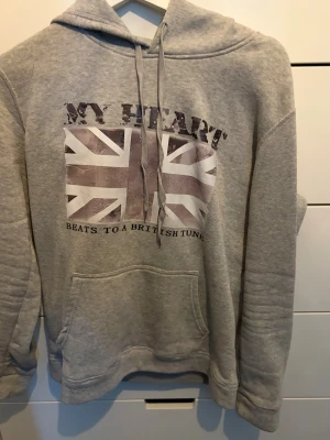 Grå hoodie med brittisk flagga - Grå hoodie med tryck av brittiska flaggan och texten 'MY HEART BEATS TO A BRITISH TUNE' pris 250