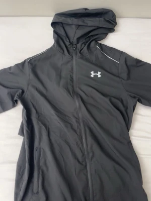 Svart vindjacka från Under Armour - Snygg svart vindjacka från Under Armour med huva och hel dragkedja framtill. Jackan har vita detaljer på axlarna och logga på bröstet samt stort tryck på ryggen. Två fickor framtill och lätt material som passar perfekt till träning eller chill. Passar även dem med storlek L.