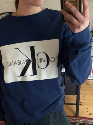 Calvin Klein Jeans sweatshirt / collegetröja navy blå med stor CK-logo - Calvin Klein Jeans sweatshirt i mörkblå/navy med stor CK-logga framtill. Snygg och klassisk collegetröja från Calvin Klein Jeans med rund hals, ribbade muddar och stor logotyp på framsidan. Modellen har en clean sportig streetwear-look och funkar perfekt till jeans, mjukisbyxor, kjol eller oversized casual outfit.  Skick: Knappt använd. Märke: Calvin Klein Jeans Färg: mörkblå/navy, vit och svart/grå logo Modell: sweatshirt / collegetröja / crewneck Storlek: M Material: 100% Bommul  