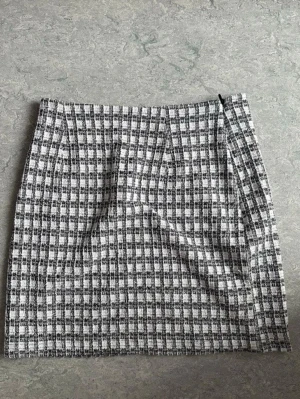 Rutig kortkjol i svart och vitt - Snygg kortkjol med rutigt mönster i svart och vitt. Kjolen har en rak passform och dragkedja i sidan. Perfekt till en enkel topp eller stickad tröja för en trendig look.