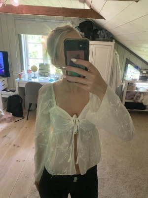 Vit blommig blus med knytning - Söt vit blus med broderade blommor. Blusen har djup urringning, knytning framtill och vida långärmade ärmar. Säljes då den ej kommer till användning och är endast använd fåtal gånger.