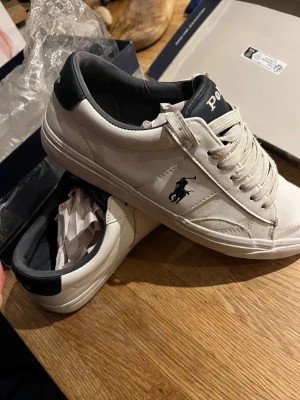 Vita Polo Ralph Lauren sneakers - Snygga vita sneakers från Polo Ralph Lauren med mörkblå detaljer och klassisk logga på sidan. Skorna har lågt skaft, vita snören och ovandel i slätt läder. Perfekta för en clean och stilren look.