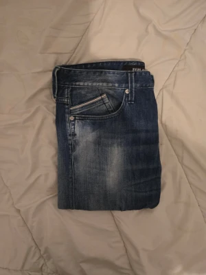 Blå jeans från Replay, w32 L32  - Säljer ett par snygga blå jeans från Replay i modell 32. Jeansen har en klassisk femficksdesign, normal midja och raka ben. Materialet är denim med slitningar och tvättade detaljer för en cool, avslappnad look. Perfekta för dig som gillar streetwear och casual stil.