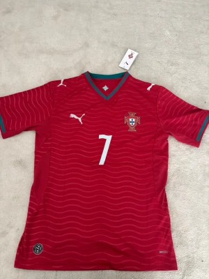 Portugal matchtröja Puma röd Storlek S - Snygg röd Portugal matchtröja från Puma med nummer 7 på bröstet och det portugisiska fotbollsförbundets emblem. Tröjan har gröna detaljer vid kragen och ärmarna, vita Puma-loggor och är tillverkad i ett lätt funktionsmaterial (DryCell). Perfekt för fotbollsfans!