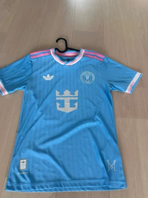 Blå Inter Miami Messi matchtröja - Snygg ljusblå Inter Miami matchtröja från Adidas med Messi och nummer 10 på ryggen. Tröjan har rosa och vita detaljer på krage och ärmslut, samt klubbmärke och Adidas-logga på bröstet. Tillverkad i lätt och ventilerande material, perfekt för fotboll eller träning.