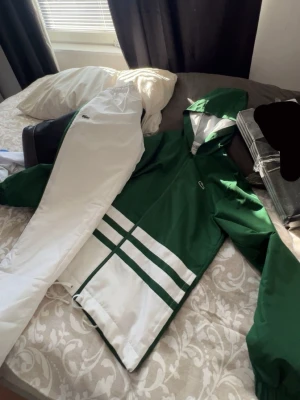 Grön och vit tracksuit från Lacoste - Snygg vindjacka från Lacoste i grönt och vitt med huva och dragkedja. Jackan har vita detaljer och två breda ränder nedtill samt den klassiska Lacoste-loggan på bröstet. Perfekt för dig som gillar sportig och stilren look.