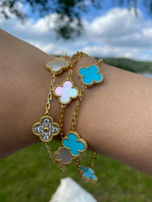 18k guldpläterad armband med snäckskal och halvädelsten  - Snyggt armband i guldton med clover-formade hängen i vitt, ljusblått och turkost. Ett av hänget är dekorerat med små pärlor och gnistrande stenar. Kedjan är tunn och elegant, perfekt för att ge din outfit en lyxig touch.