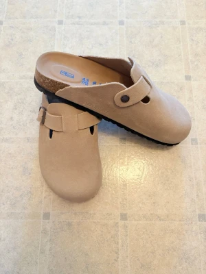 Beige Birkenstock tofflor i mocka - Säljer ett par beigea Birkenstock tofflor i mocka med klassisk design och justerbart spänne. De har en bekväm korksula och svart räfflad yttersula för bra grepp. Perfekta för dig som gillar stilrena och sköna skor. Hör av dig vid frågor eller andra funderingar!🙂