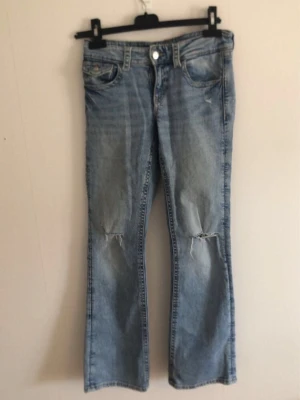 Ljusblå bootcut jeans med slitningar - Säljer ett par ljusblå bootcut jeans med slitningar över knäna och klassiska fem fickor. Jeansen har en avslappnad passform och är tillverkade i denim. Perfekta för en cool och avslappnad stil.