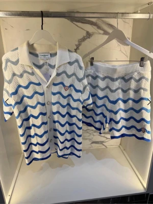 Vit och blå Casablanca shortset - Säljer ett snyggt set från Casablanca med kortärmad skjorta och shorts i virkad stil. Setet är vitt med vågmönster i blått och grått, har krage och knappar framtill samt ribbade kanter. Perfekt för dig som gillar trendiga och bekväma plagg med unik design.