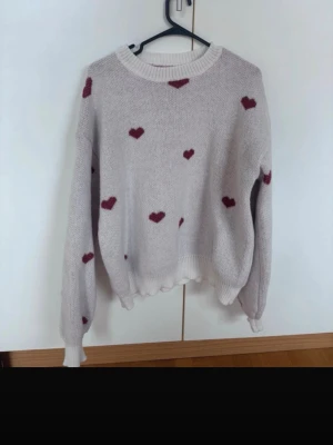 Beige stickad tröja med hjärtan - Söt beige stickad tröja med röda hjärtmönster över hela tröjan. Tröjan har rund halsringning, långa ärmar och ribbade muddar vid ärmslut och nederkant. Perfekt för dig som gillar gulliga detaljer och en avslappnad stil.