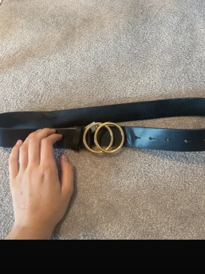 Svart bälte med guldfärgat spänne - Snyggt svart bälte med dubbla guldfärgade ringar som spänne. Bältet har en stilren design och passar perfekt till jeans eller kjol. Tillverkat i skinnimitation med flera hål för justerbar passform.