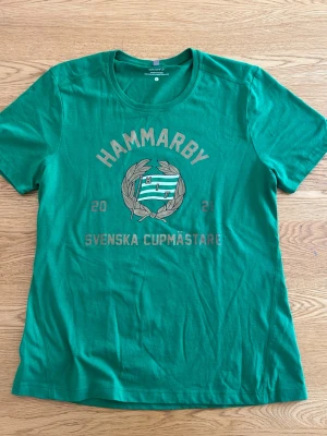 Grön Hammarby t-shirt Cupmästare - Grön t-shirt från Craft med tryck 'Hammarby Svenska Cupmästare 2021' och klubbens emblem på bröstet. Klassisk rund hals och korta ärmar. Perfekt för dig som vill visa ditt stöd för Hammarby på ett stilrent sätt. Det är storlek L, men sitter som en S/M