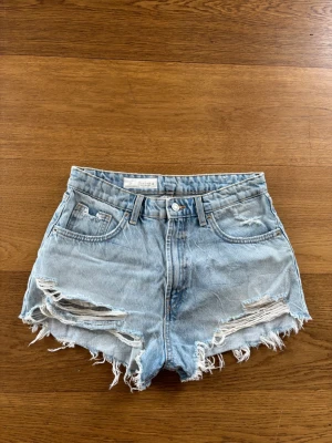 Ljusa denimshorts från ZARA - Säljer ett par ljusblå denimshorts från ZARA med hög midja och slitna detaljer. Shortsen har fransig kant, klassiska fem fickor och knappgylf. Perfekta för sommaren och ger en avslappnad vibe.