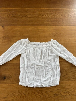 Vit offshoulder blus med långa ärmar subdued  - Säljer en vit offshoulder blus med långa ärmar och smockad resår upptill. Blusen har en luftig och lätt känsla, perfekt för en avslappnad och trendig look. Materialet känns mjukt och följsamt, och den har en lös passform.