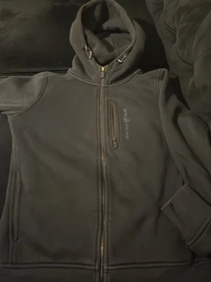 Mörkgrå hoodie från Peak Performance - Säljer en mörkgrå hoodie från Peak Performance med dragkedja framtill och en bröstficka med zip. Hoodien har justerbar huva med snören och diskret logga på bröstet. Perfekt för chill dagar eller när du vill ha något bekvämt och stilrent.
