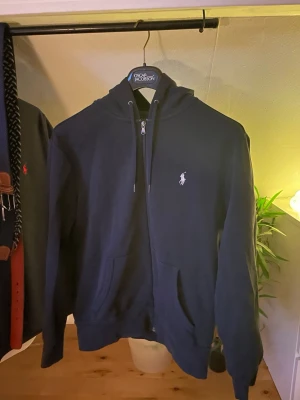 Marinblå hoodie från Ralph Lauren - Snygg marinblå hoodie från Ralph Lauren med dragkedja framtill, två fickor och klassisk vit broderad logga på bröstet. Hoodien har huva med dragsko och ribbade muddar vid ärmslut och nederkant. Perfekt för en avslappnad och stilren look.